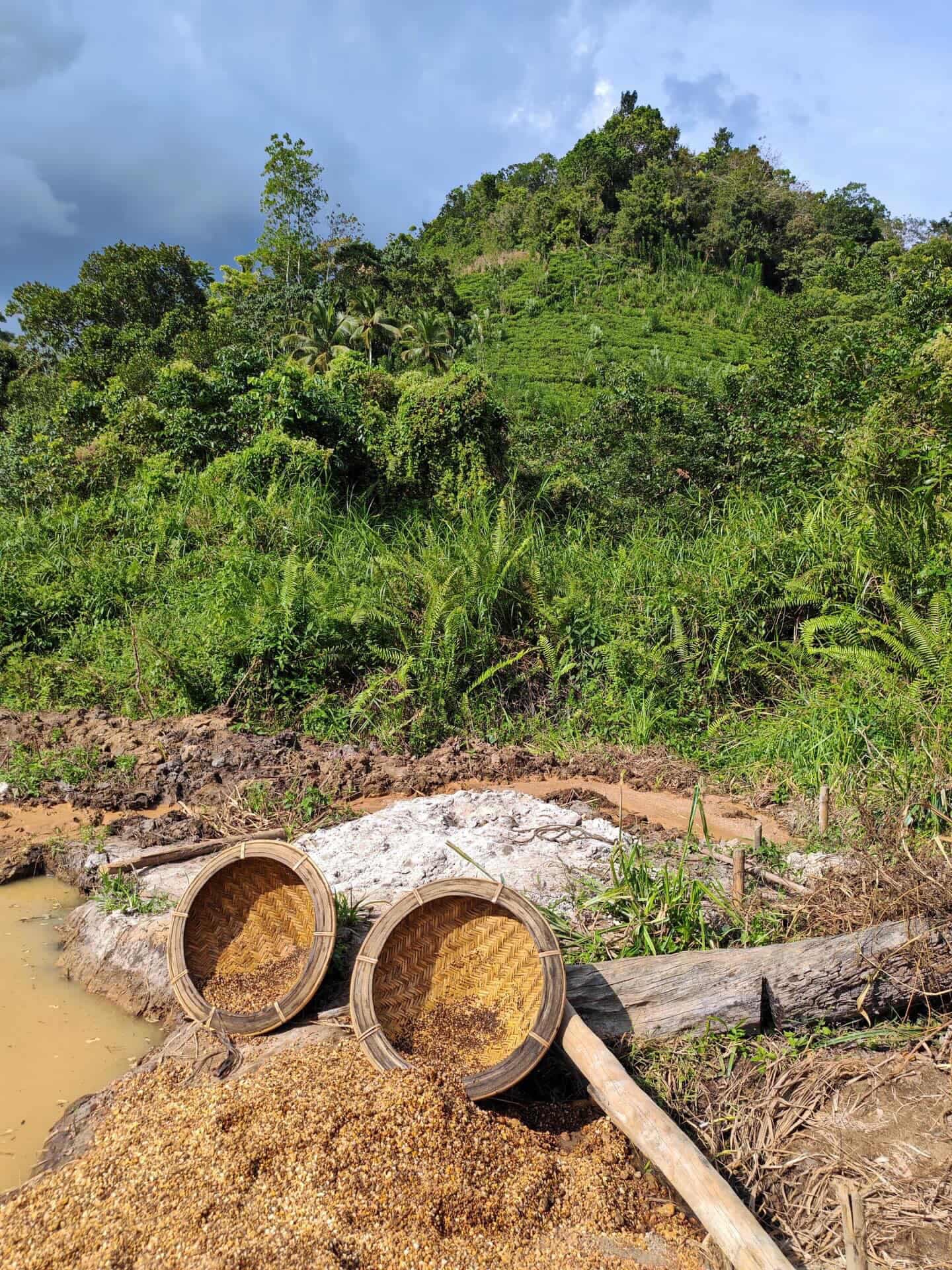 Two woven baskets of washed gravel at the edge of a gem pit, a forested hillside rising behind them